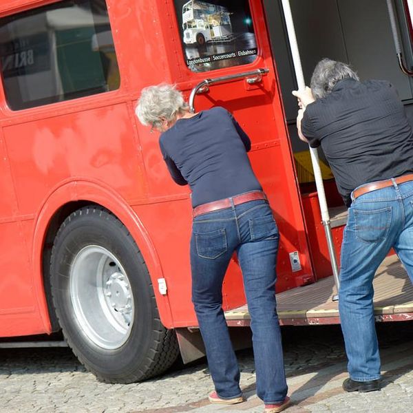 Zwei Personen schieben gemeinsam einen roten Bus.