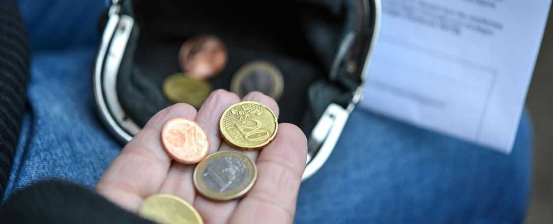 Eine offene Handfläche, auf der mehrere Euro-Münzen liegen, über einer schwarzen Geldbörse. Im Hintergrund liegt eine Rechnung.
